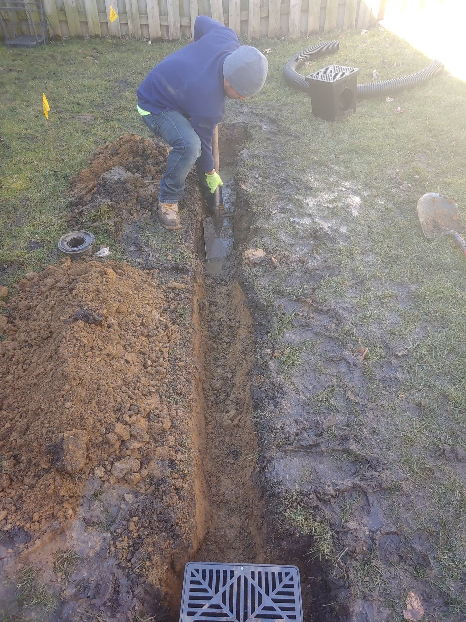 Worker setting drainage pipe in a muddy trench with a square catch-basin grate in the foreground