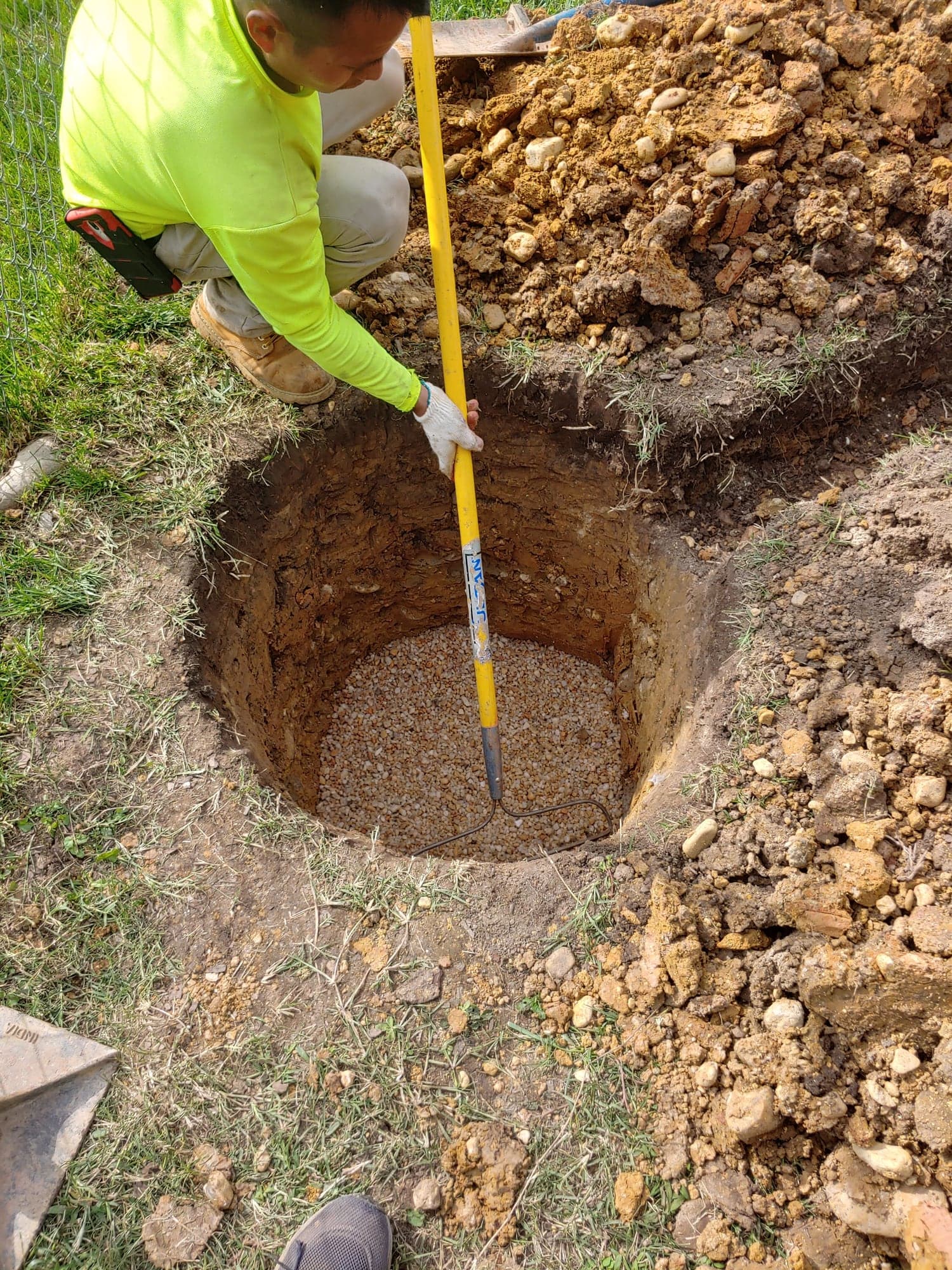 Worker with a shovel beside a deep round dry-well pit bedded with clean gravel