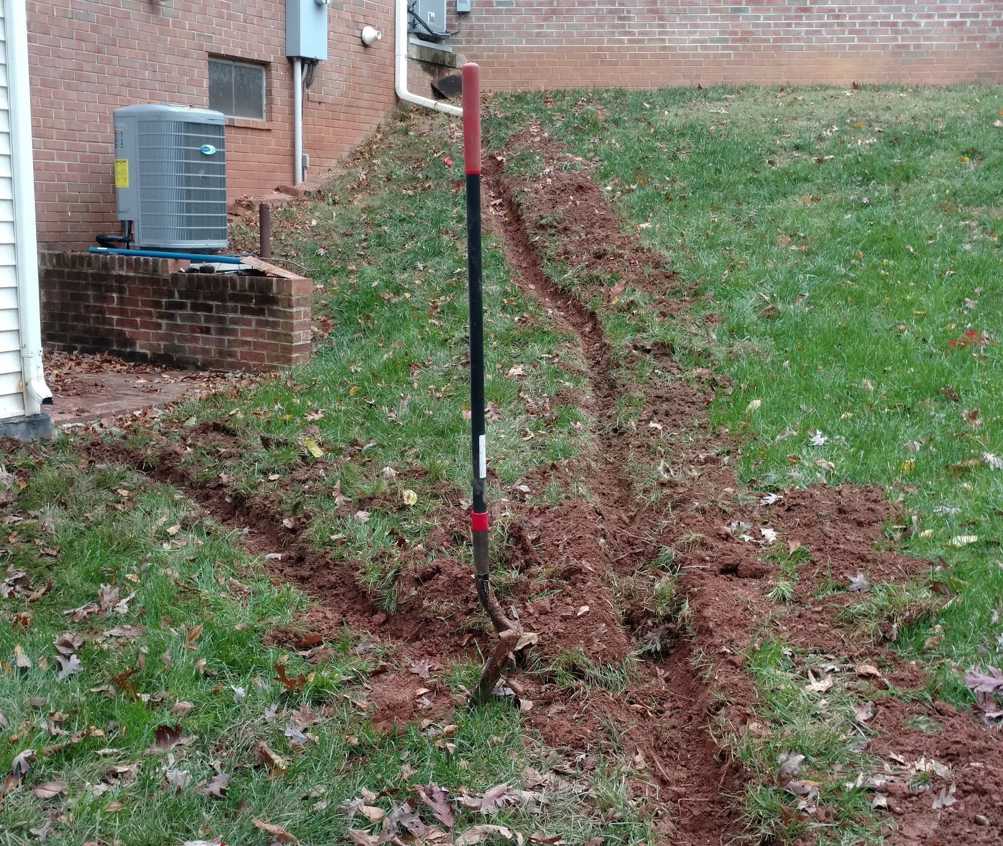 Hand-dug drainage trench cut along a grassy slope next to a brick house and HVAC pad