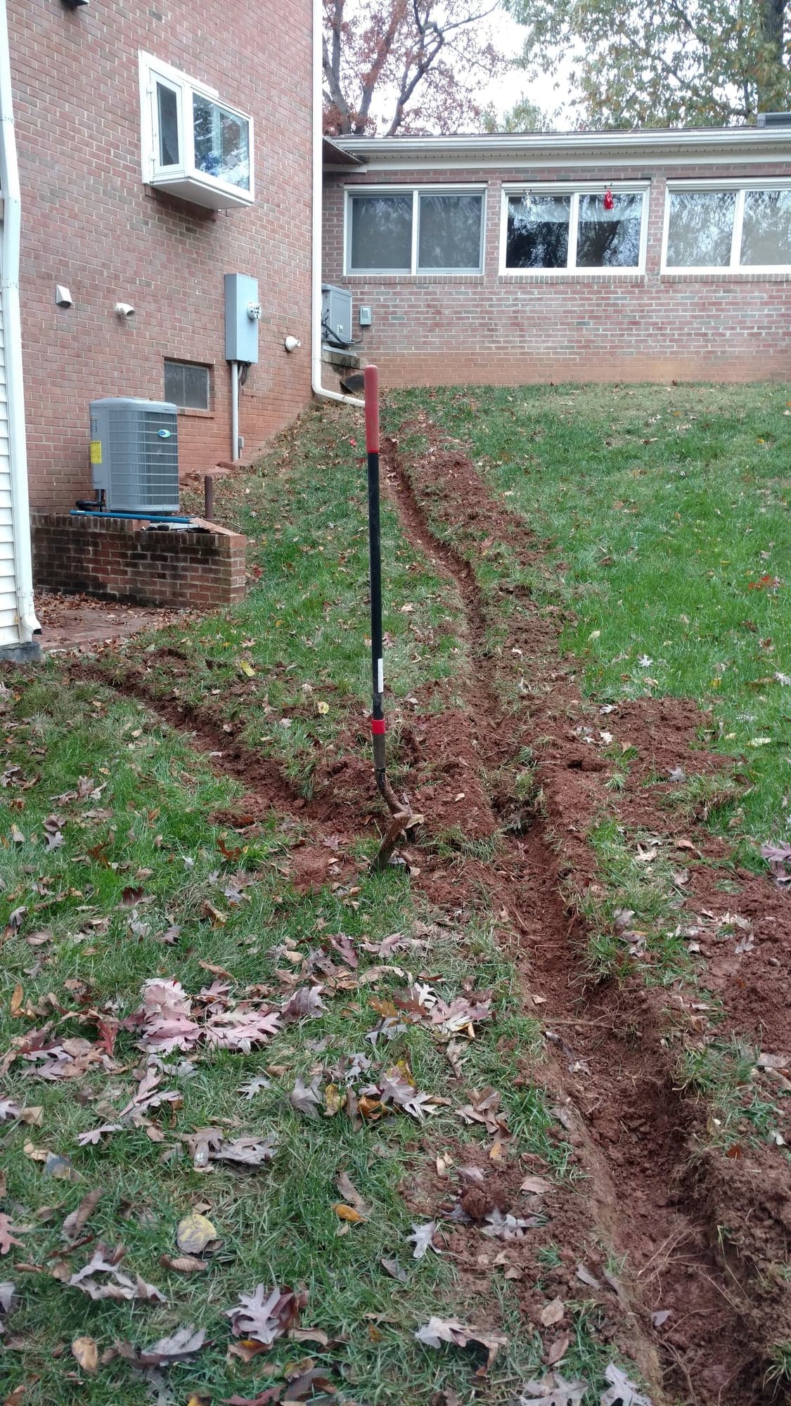 Narrow hand-dug trench running down a grassy slope beside a brick house and its air-conditioning unit