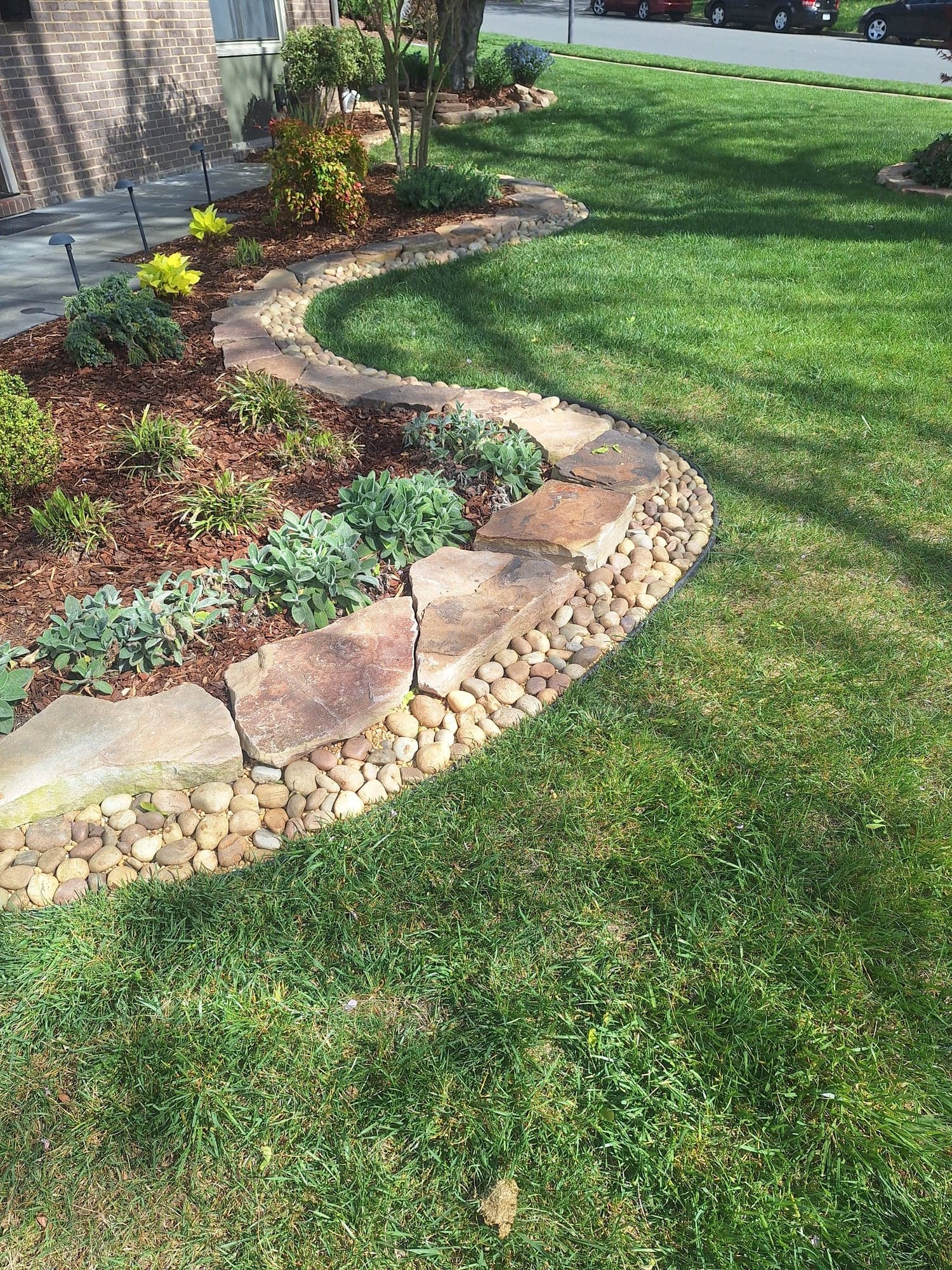 Curved planting bed with a flagstone wall and river-rock edging along a green lawn